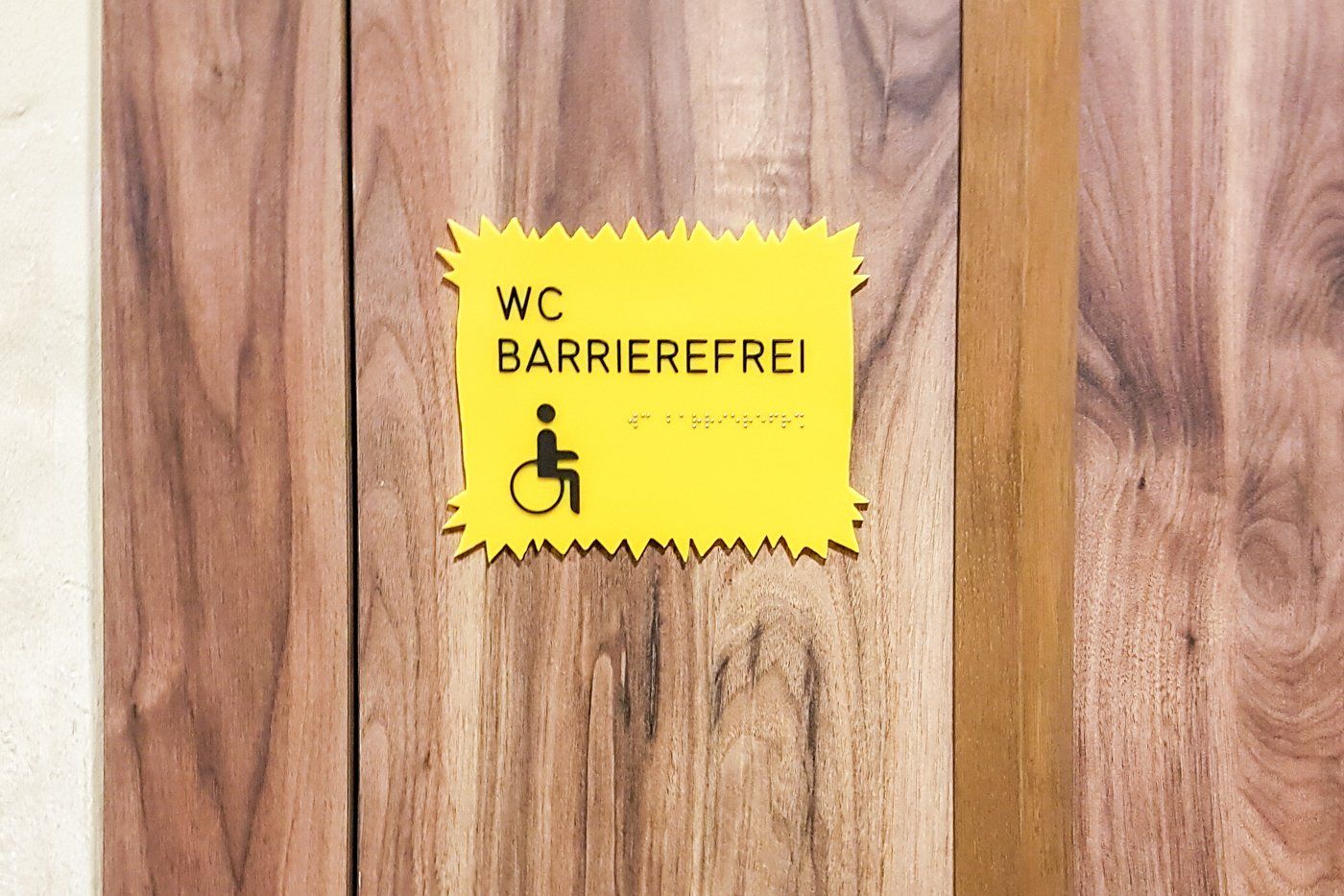 Auffälliges neongelbes Schild mit gezackten oberen und unteren Kanten, deutlich gekennzeichnet als barrierefreies WC. Der Text 'WC barrierefrei' ist sowohl in konventioneller Schrift als auch in taktilem Braille für Sehbehinderte hervorgehoben. Unterhalb des Schriftzugs ist das universell erkennbare Symbol einer Figur im Rollstuhl abgebildet, was die Zugänglichkeit für Rollstuhlfahrer betont.