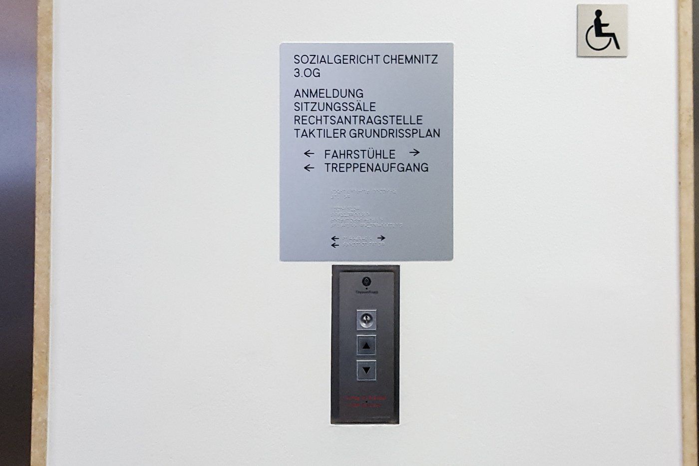 Ein taktiles Schild des Sozialgericht Chemnitz. Darunter befinden sich Aufzugknöpfe. Das Schild beschreibt, wo die Fahrstühle und Treppen zu finden sind.