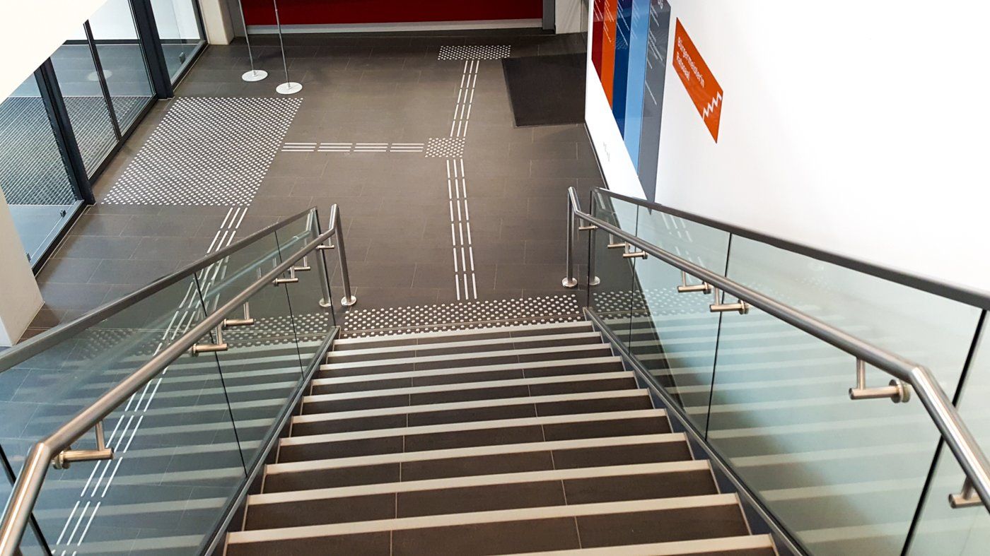 Eine Treppe mit gläsernem Treppengeländer und mit weißen Stufenmarkierungen führt in den Eingangsbereich. Der Ausgang kann von der Treppe über ein Bodenleitsystem erreicht werden.