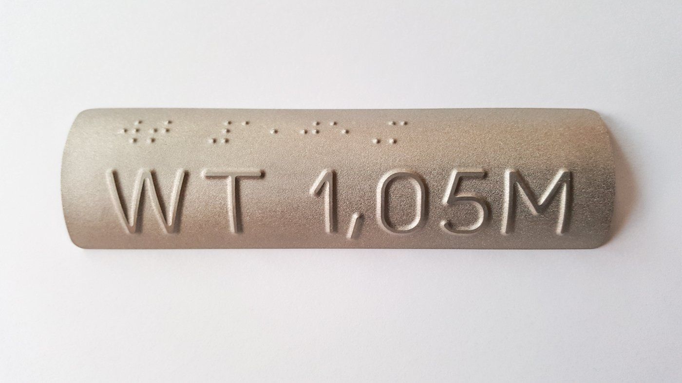 Ein taktiles Handlaufschild eus Edelstahl mit der Aufschrift WT 1,05M. Die lateinischen Buchstaben sind oberhalb in Brailleschrift übersetzt.