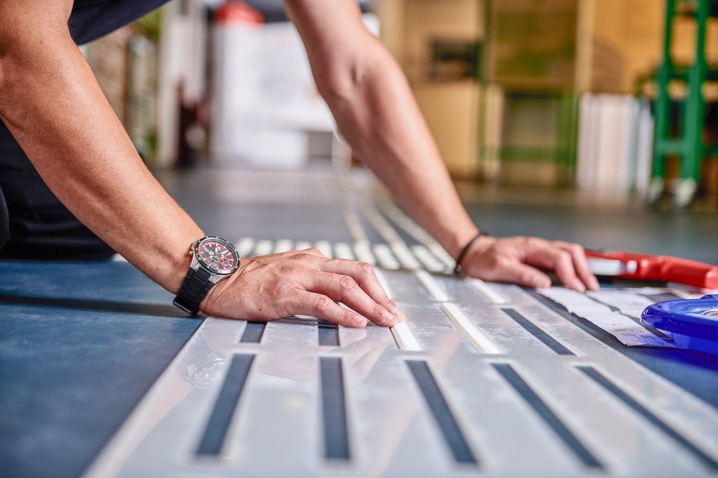 Hände richten ein Bodenleitsystem mit einer Schablone auf einem Boden aus.  Die linke Hand liegt mit dem Ballen auf einem Streifen auf. Die rechte Hand drückt einen Streifen mit dem Finger fest.