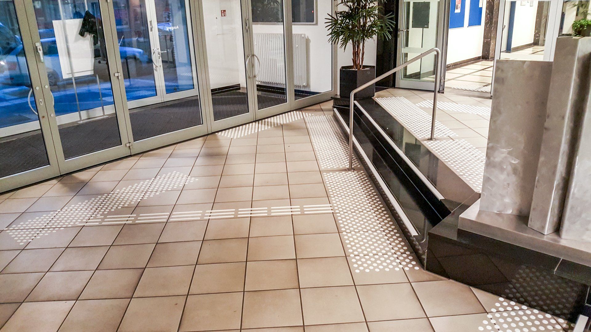 Eingangsbereich mit Glastüren und Bodenleitsystem und Treppenmarkierungen