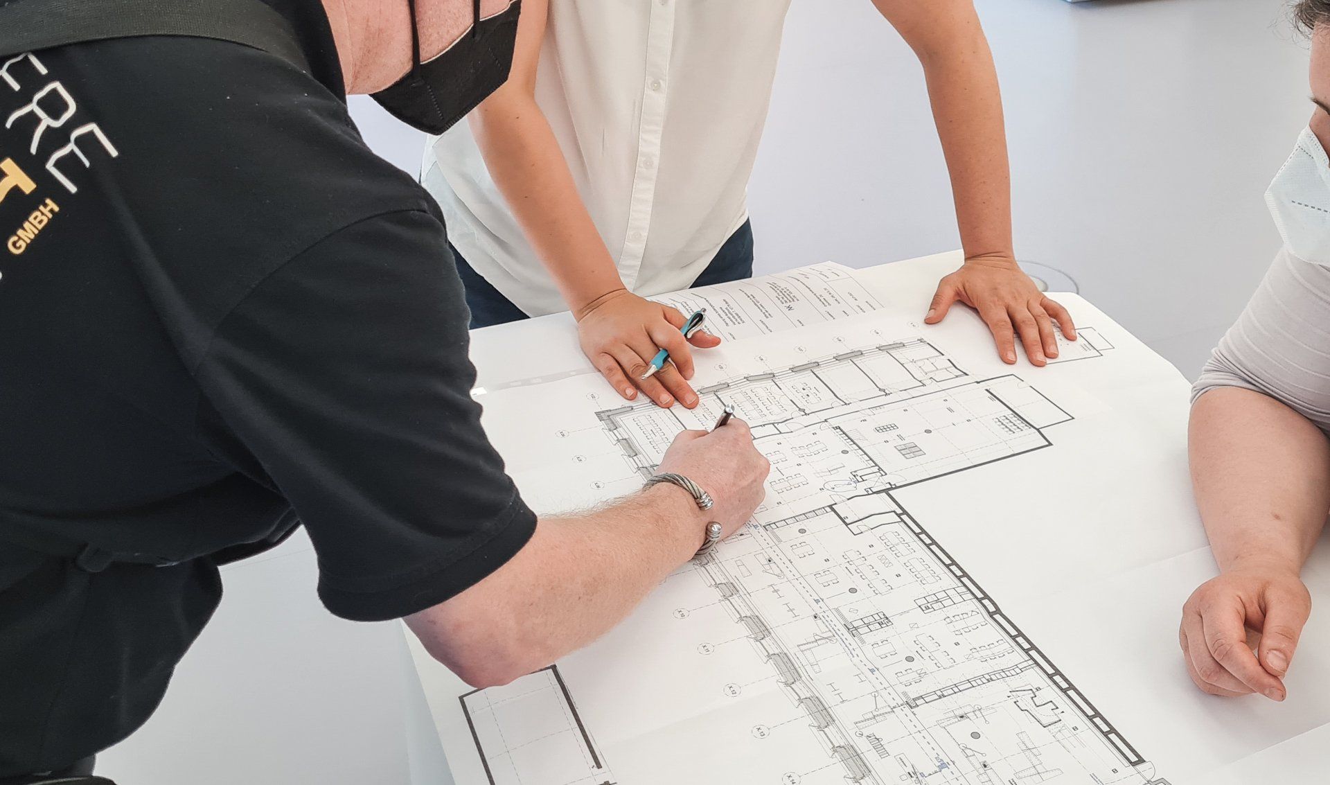 Unser Mitarbeiter berät über den Grundriss eines Gebäudes gelehnt zwei Kundinnen in Bezug auf die barrierefreie Wegeführung und der Montage eines visuellen und taktilen Leitsystems.