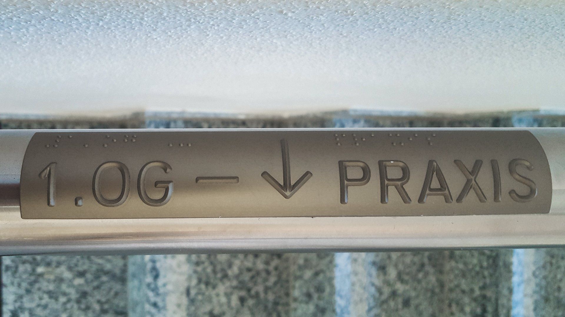 Handlaufschild mit der Aufschrift 1.OG und einem Pfeil nach unten, neben dem Praxis steht