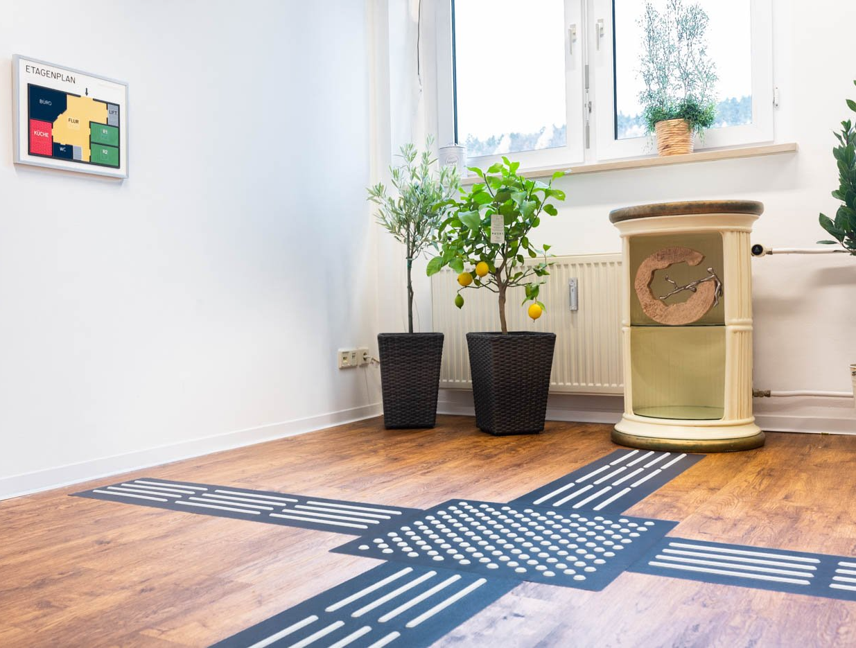 Rippenplatten und ein Aufmerksamkeitsfeld sind auf einem Boden verlegt. Das Aufmerksamkeitsfeld leitet in vier Richtungen weiter. Man kann so zu einem Etagenplan gehen oder einem Beistelltisch kommen.