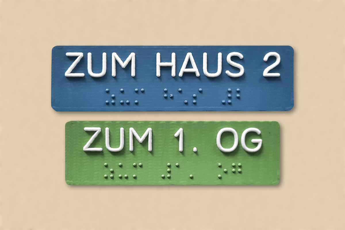 Ein grünes und ein blaues taktiles Handlaufschild an einer Wand mit den Aufschriften zum Haus 2 und zum 1. OG. Die lateinischen Buchstaben sind unterhalb in Brailleschrift übersetzt.