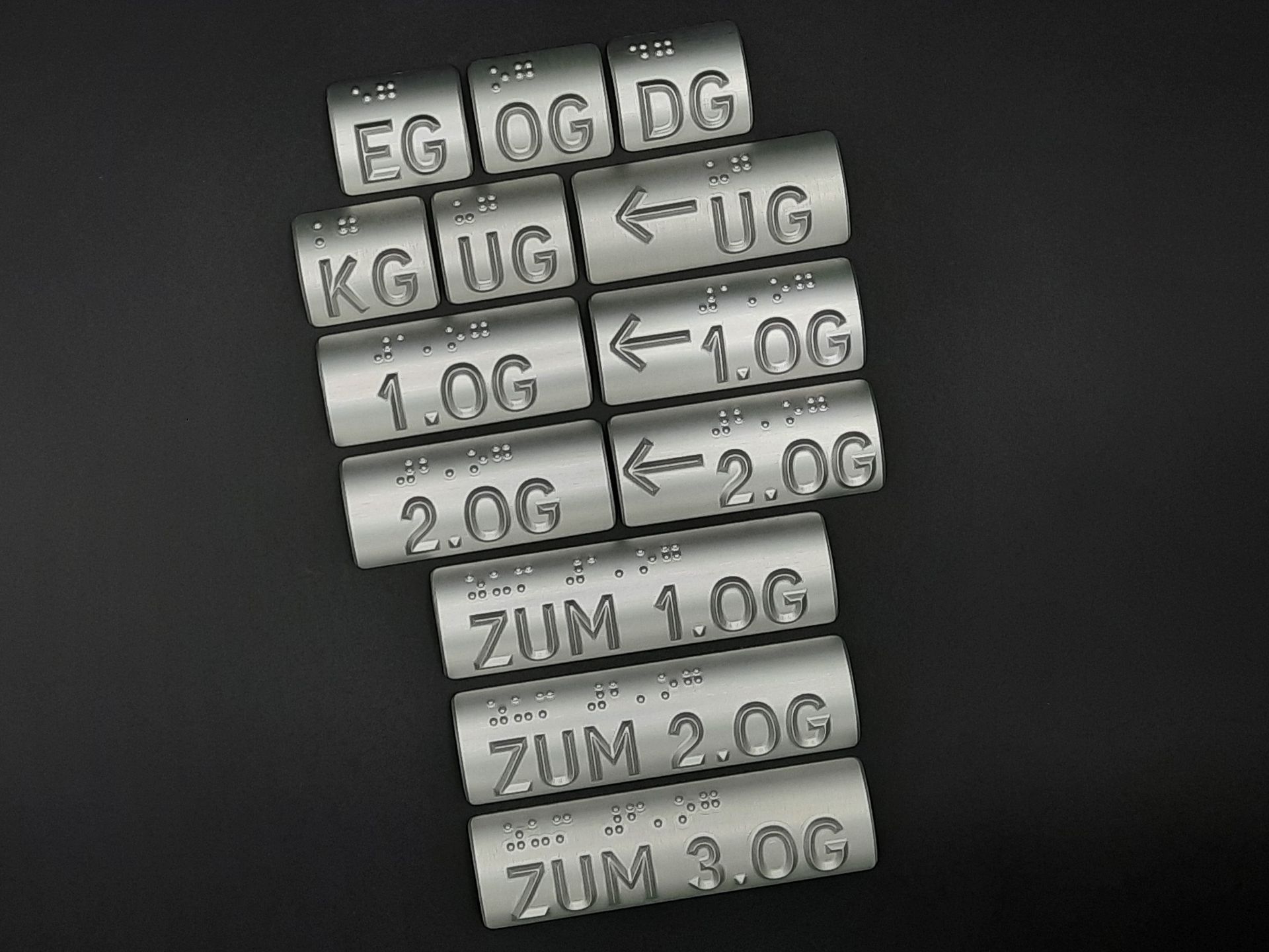 Silberne Handlaufschilder, die beinhalten, welches Stockwerk die aktuelle Etage ist und welche Etage über den Handlauf erreicht werden kann. Die lateinischen Buchstaben sind oberhalb in Brailleschrift übersetzt.