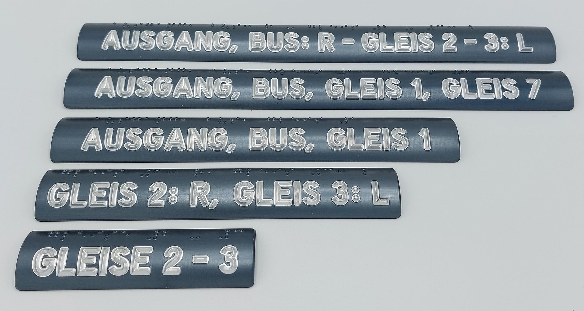 Dunkelgraue Handlaufschilder aus Aluminium. Auf den Schildern steht, welches Gleis über den Handlauf erreicht werden kann. Die lateinischen Buchstaben sind oberhalb in Brailleschrift übersetzt.
