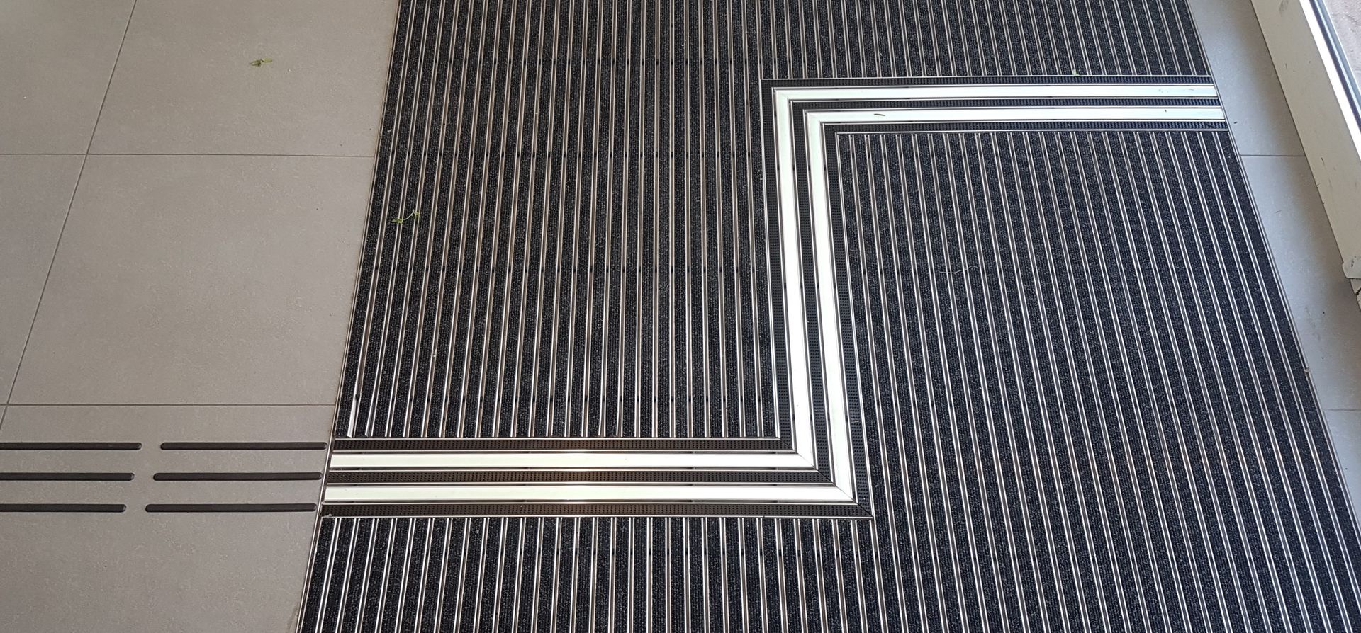 Eine Sauberlaufmatte vor einer Eingangstür. Die Leitspur führt Besucher nicht über geraden Wege, sondern über zwei Ecken in die Innenräume. Die Spur und die Ecken sind in die Matte integriert.