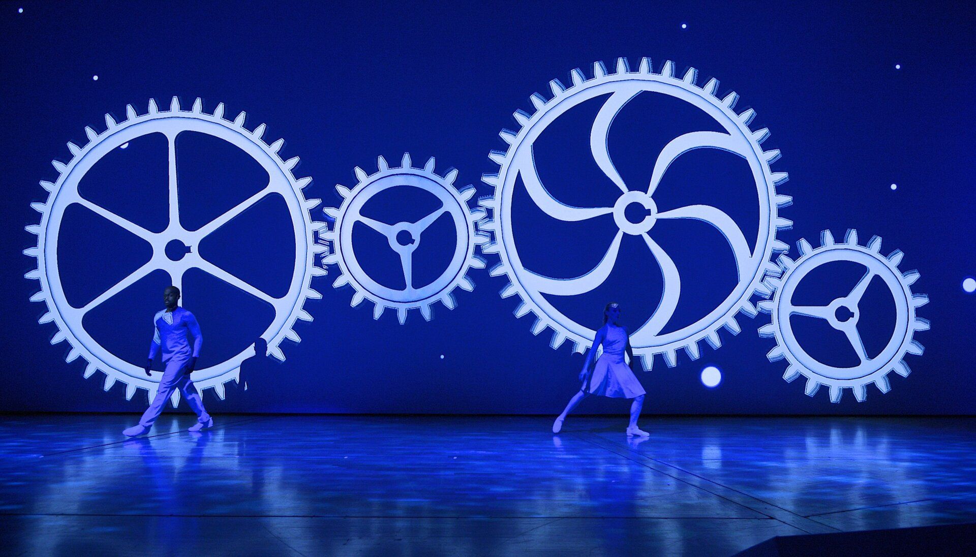 Zwei Tänzer auf einer blauen Bühne vor riesigen Zahnrädern
