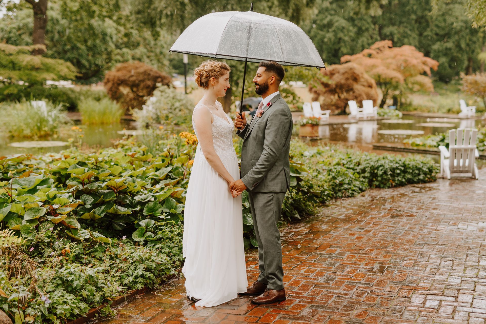 Es regnet, das Brautpaar steht steht unter einem Regenschirm,sie blicken sich verliebt an. Er trägt einen salbeifarbenen Anzug und sie trägt ein fließendes Kleid mit Spitzenoberteil.Ihre blonden Haare sind locker aufgesteckt .