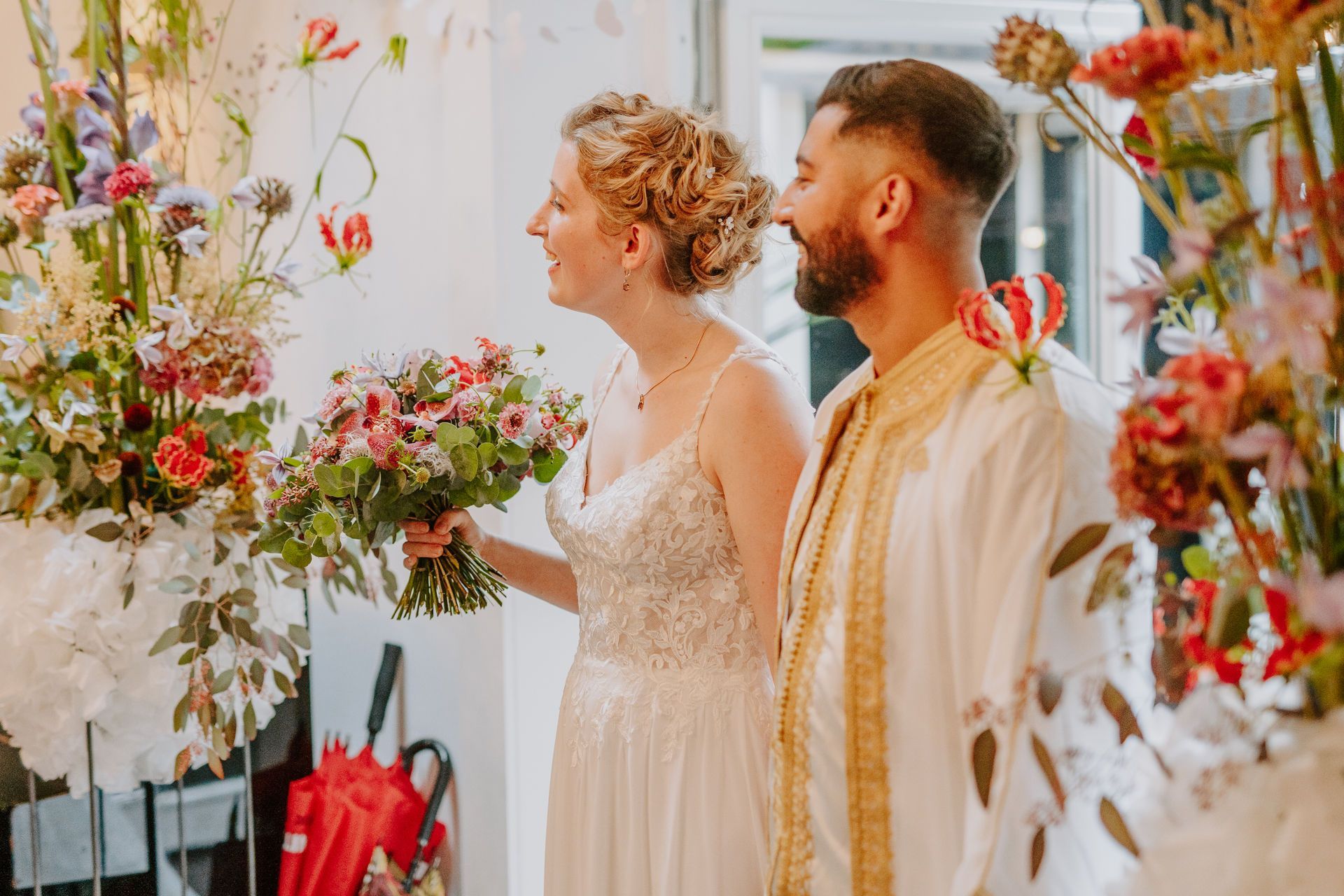 Brautpaar inmitten von leuchtenden Blumen feiern ihre moderne Oriental Boho Hochzeit.Der Bräutigam trägt eine Dishdasha, die Braut ein Bohokleid. Ihre blonden Haare sind hochgesteckt.