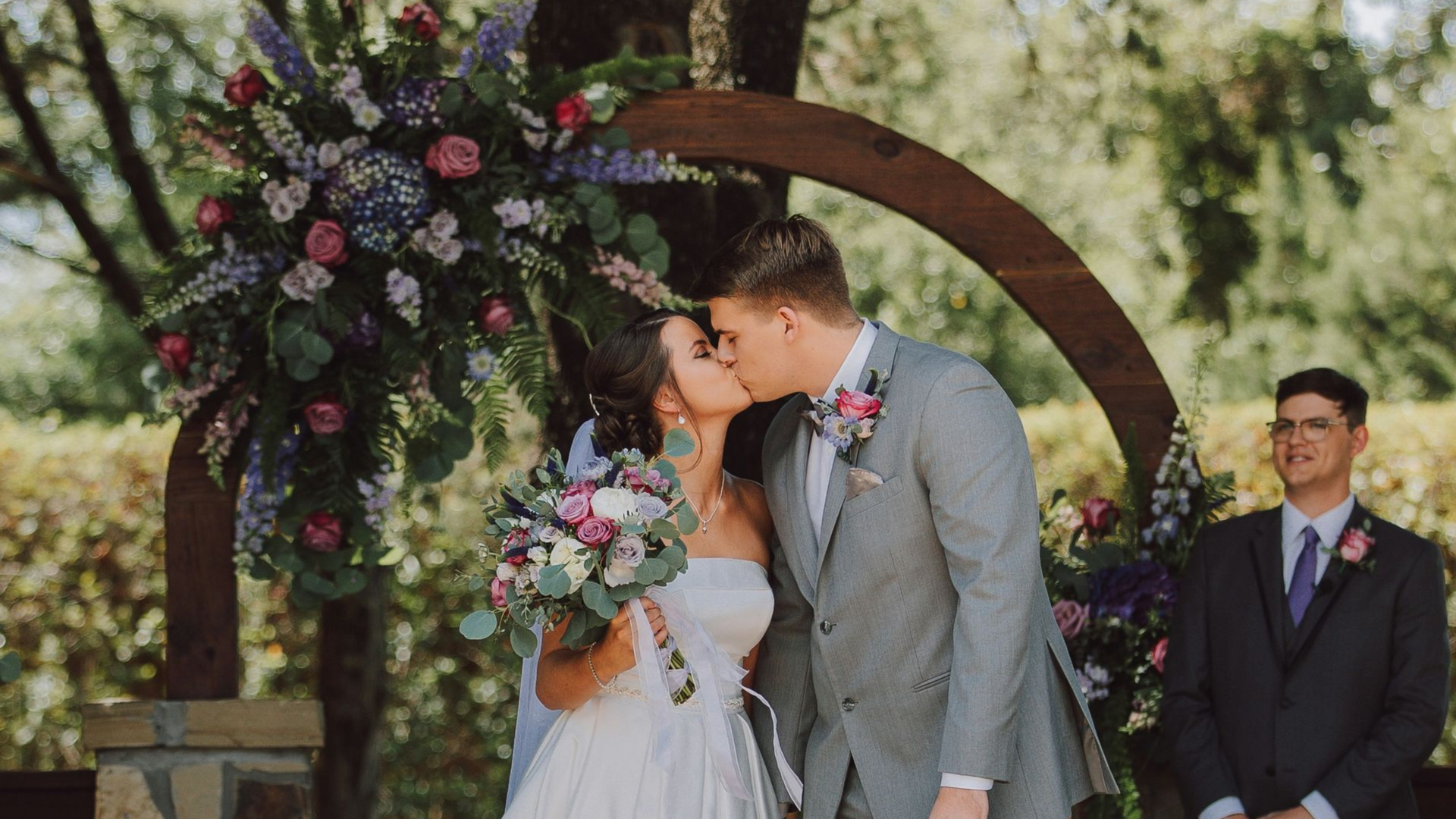 Brautpaar küsst sich unter Traubogen aus Holz vor dem Trauredner im Hintergrund Brautstrauß mit Blumen in lila rosa grün weiß | Fotocredit: Nate Johnston / Unsplash