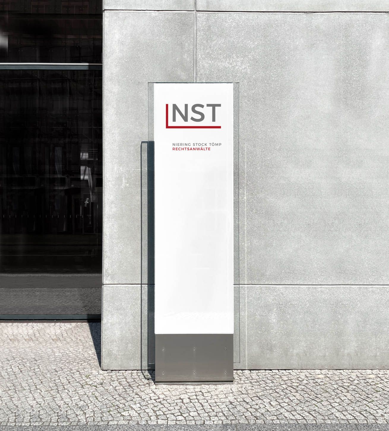 NST-Wandstele NST-Wandstele mit NST-Logo im Außenbereich vor einer grauen Betonwand und Kopfsteinpflaster.