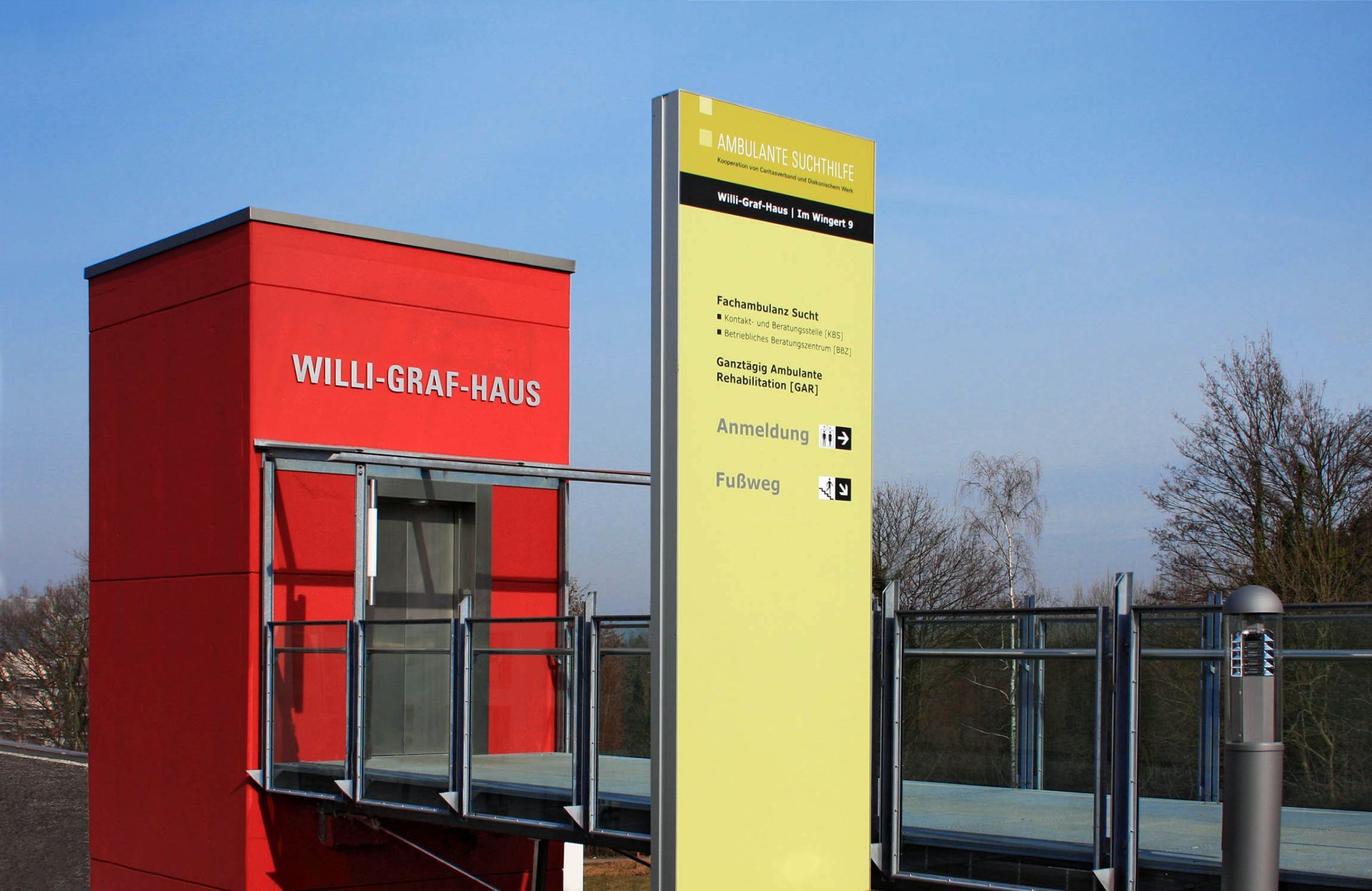 Eingang mit Infostele Eingang des roten Aufzugturms zum Willi-Graf-Haus mit gelbgrüner Stele im Vordergrund.