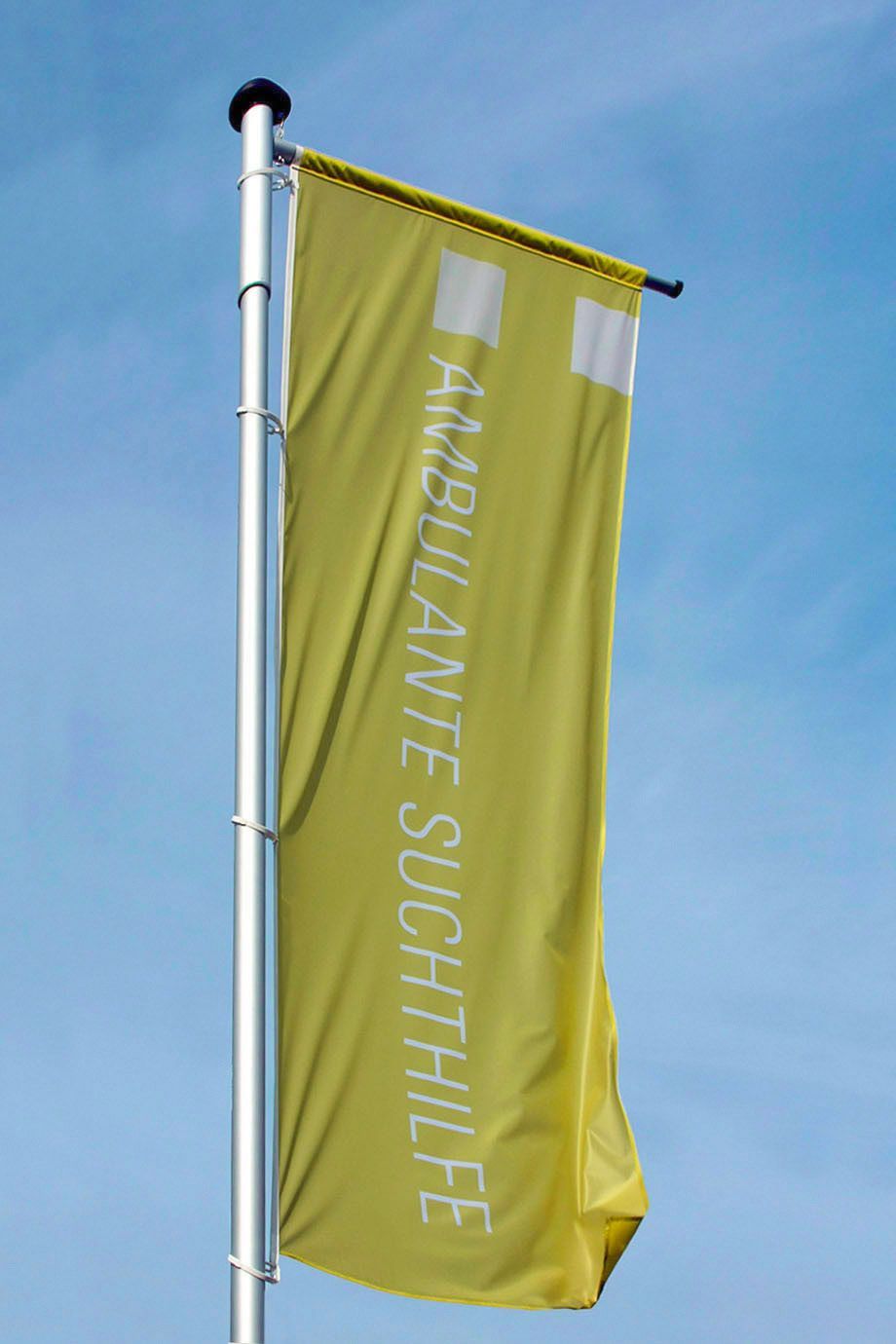 Fahne Fahne an Metallstange vor blauem Himmel mit Logoschriftzug der ambulanten Suchthilfe Bonn und auf gelbgrüner Fläche der Corporate-Designfarbe.