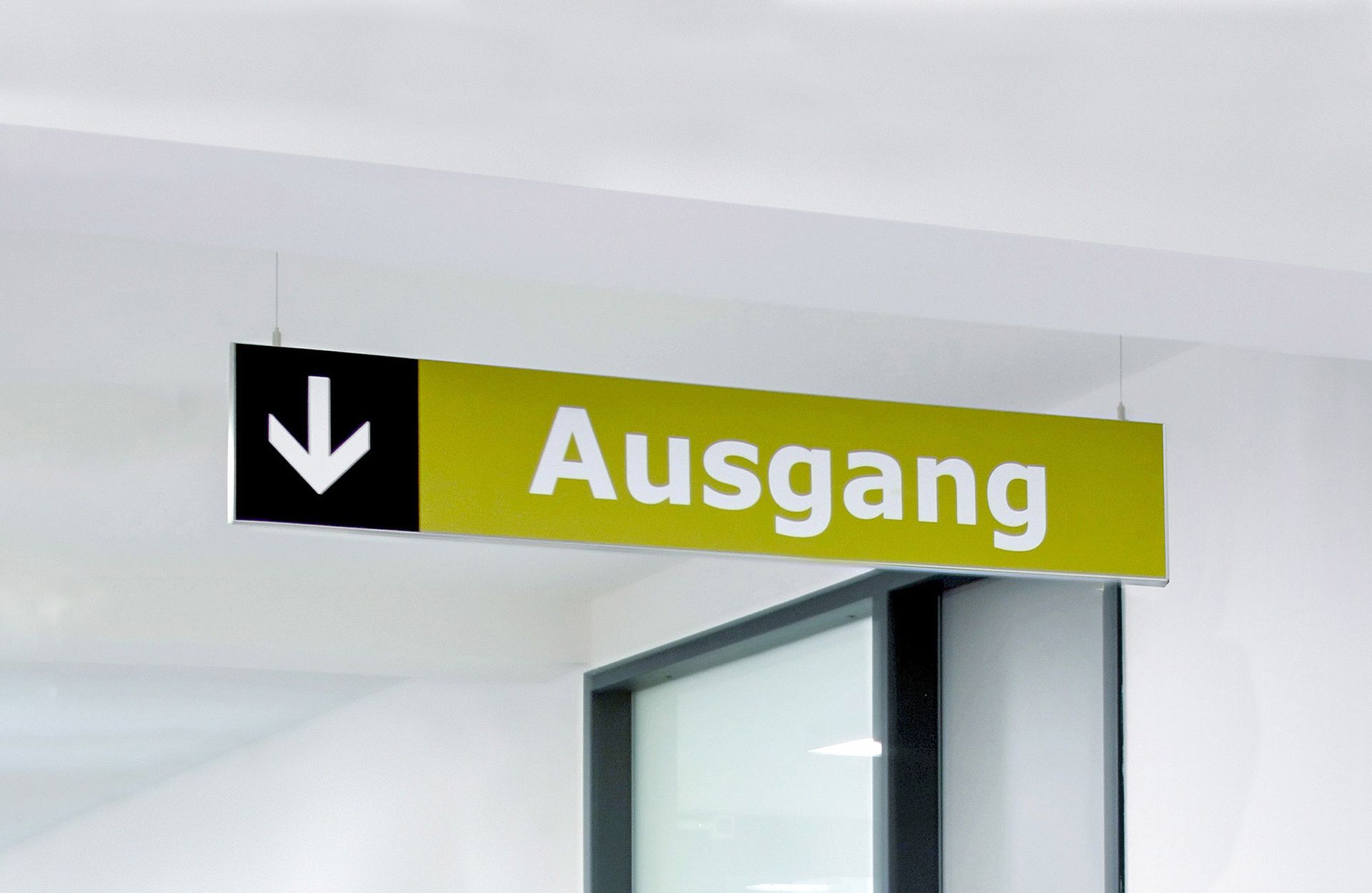 Ausgangsschild Ein schmales Ausgangsschild hängt von der Decke. Ein weißer Pfeil im schwarzen Kasten sowie der Hinweis  „Ausgang“ in weißer Farbe auf gelbgrünen Hintergrund ist auf dem Schild zu sehen.