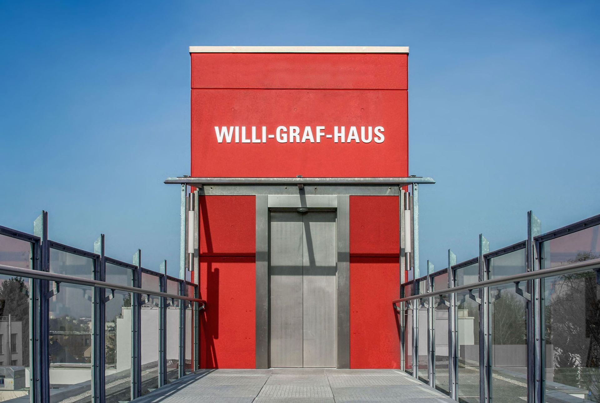Eingang Aufzug zum Willi-Graf-Haus Eingang zum Willi-Graf-Haus in Bonn über die Tür eines roten Aufzugsturms. Der Name ist als weißer Schriftzug über der metallischen Schiebetür auf der roten Wand angebracht. Ein Geländer aus Glas und Metall führt perspektivisch zur Eingangstüre.