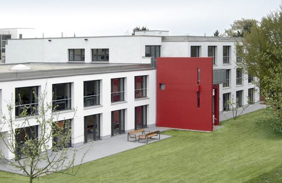 Willi-Graf-Haus Seitenansicht Seitenansicht des Willi-Graf-Hauses mit grüner Wiese und Garten. Das Haus hat eine weiße Fassade und wird in der Mitte von einer roten Betonwand optisch getrennt.