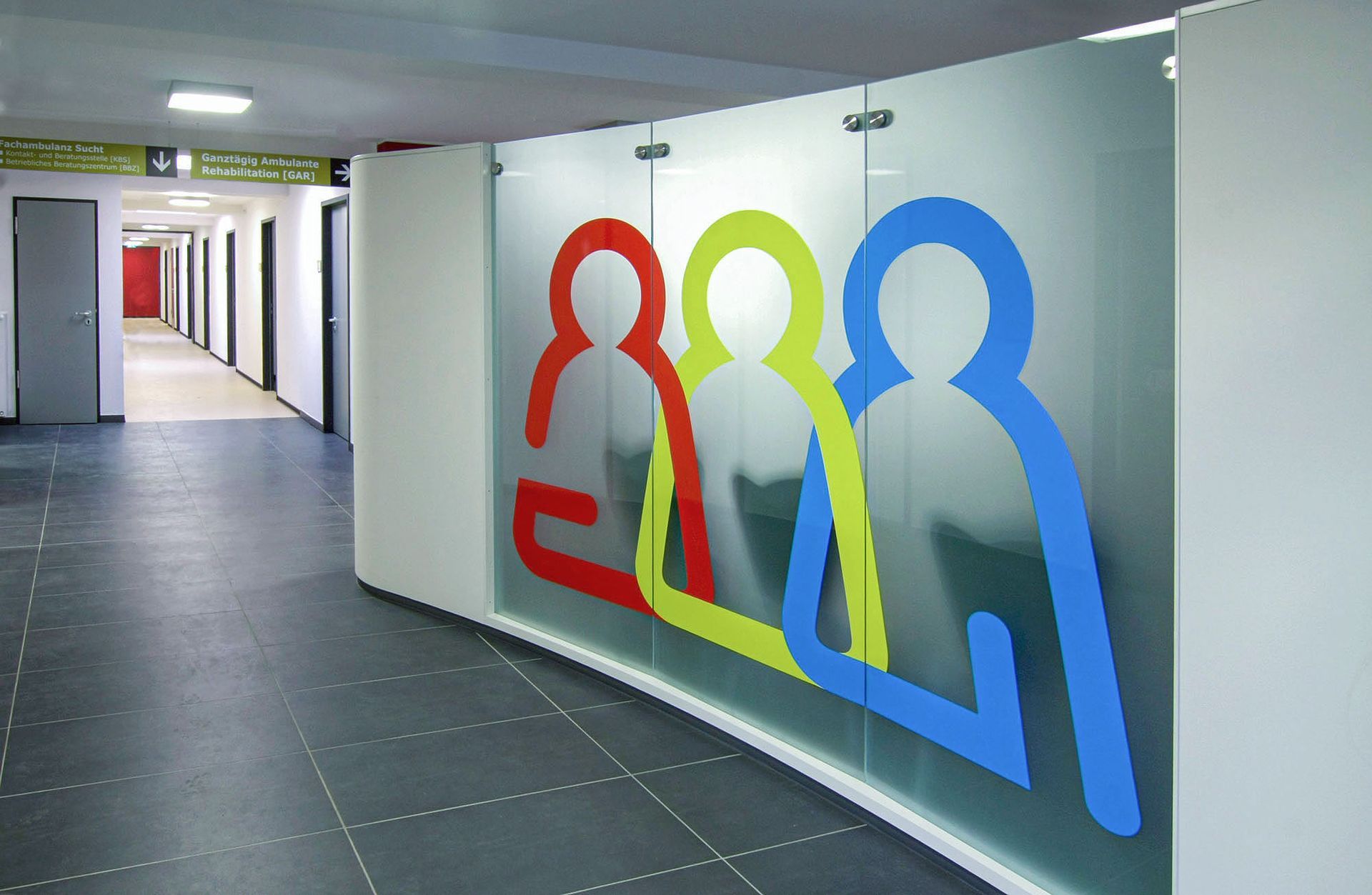 Wartebereich Wartebereich im Willi-Graf-Haus mit einer Trennwand aus Glas. Eine Illustration aus drei stilisierte Personen sind im Iconstil und in den Farben Rot, Gelbgrün und Blau sind über die ganze Fläche der Glaswand angebracht.