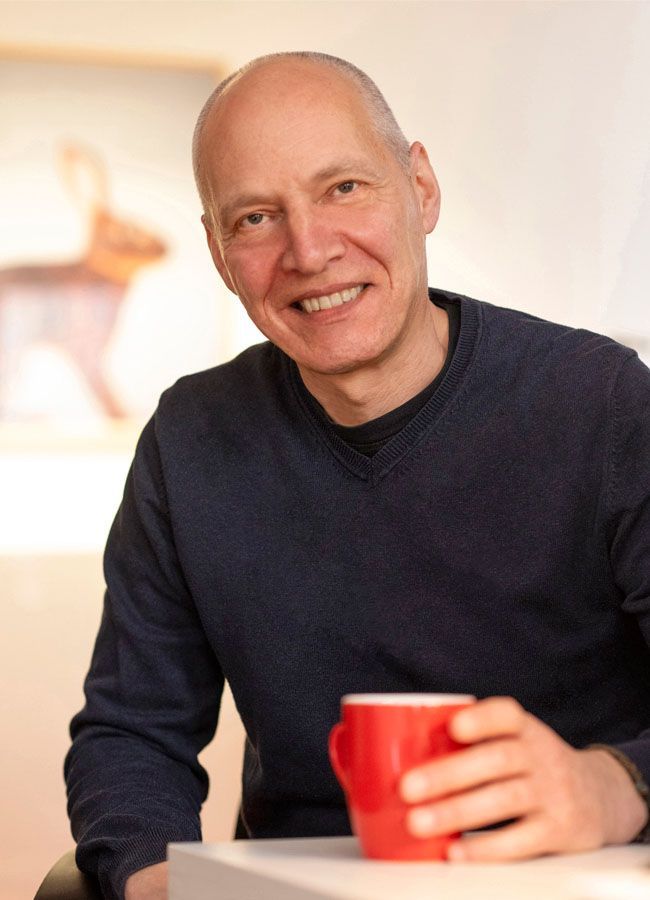 Wolfgang Gast Wolfgang Gast, Portraitfoto, Blickrichtung zum Bildbetrachter, sitzend am Tisch, der Oberkörper ist zu sehen, dunkler Pullover, leicht unscharfe, rote Kaffeetasse stehe im Vordergrund und wird von der linken Hand festgehalten