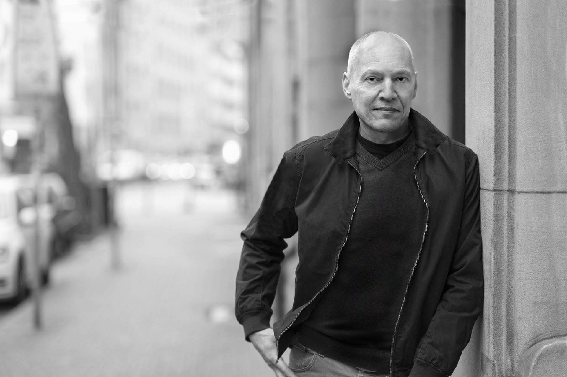 Wolfgang Gast Portraitfoto von Wolfgang Gast, schwarzweiss, Querformat, Blickkontakt zum Betrachter, an einer Hausfassade aus Sandstein angelehnt, Gesicht und Oberkörper zum Betrachter zugewandt, mit Sommerjacke und Pullover. Die linke Bildhälfte zeigt eine Straßenflucht, die in die eine Unschärfe führt.