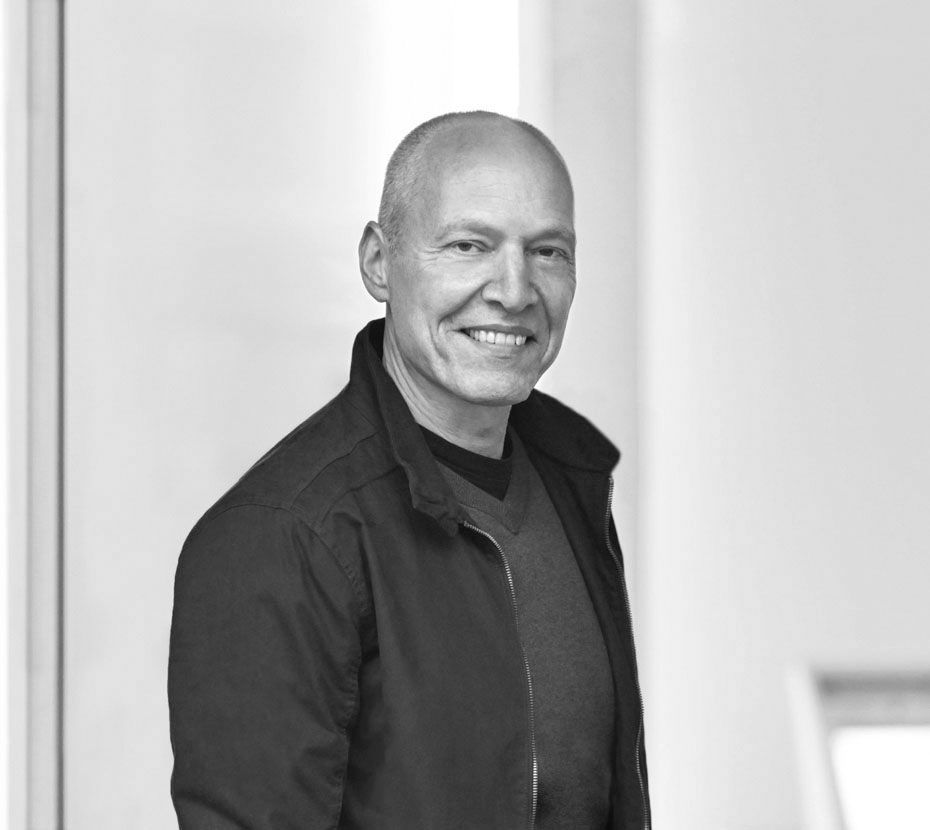 Wolfgang Gast Portraitfoto in schwarzweiss von Wolfgang Gast, mit dunkler geöffneter Sommerjacke und Pullover, Blickkontakt in die Kamera und zum Betrachter, Der Körper ist bis zum Brustkorb zu sehen, der Hintergrund ist hell und unscharf