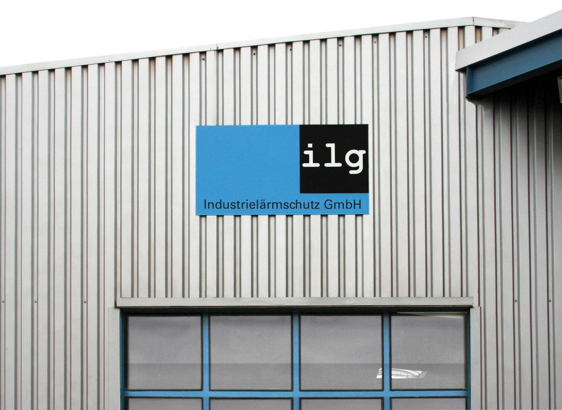 Hallenwand Fassade einer Werkshalle aus Blech und Eingangstor im unteren Bildanschnitt. Darüber ist ein Schild angebracht, das aus blaugrauem Hintergrund und dem ilg-Logo besteht. Unter dem Logo steht auf ganzer Schildbreite: „Industrielärmschutz GmbH“.