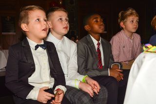 Kinder sitzen auf ein Objekt blickend. Kinder in festlicher Kleidung blicken auf eine - auf dem Foto nicht sichtbare - Leinwand.