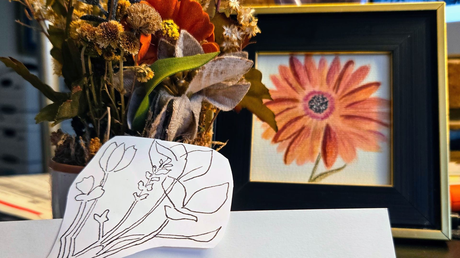 Picture shows a mixed boquet with dried flower stems, a black and white sticker with a botanical drawing, and a framed floral painting.