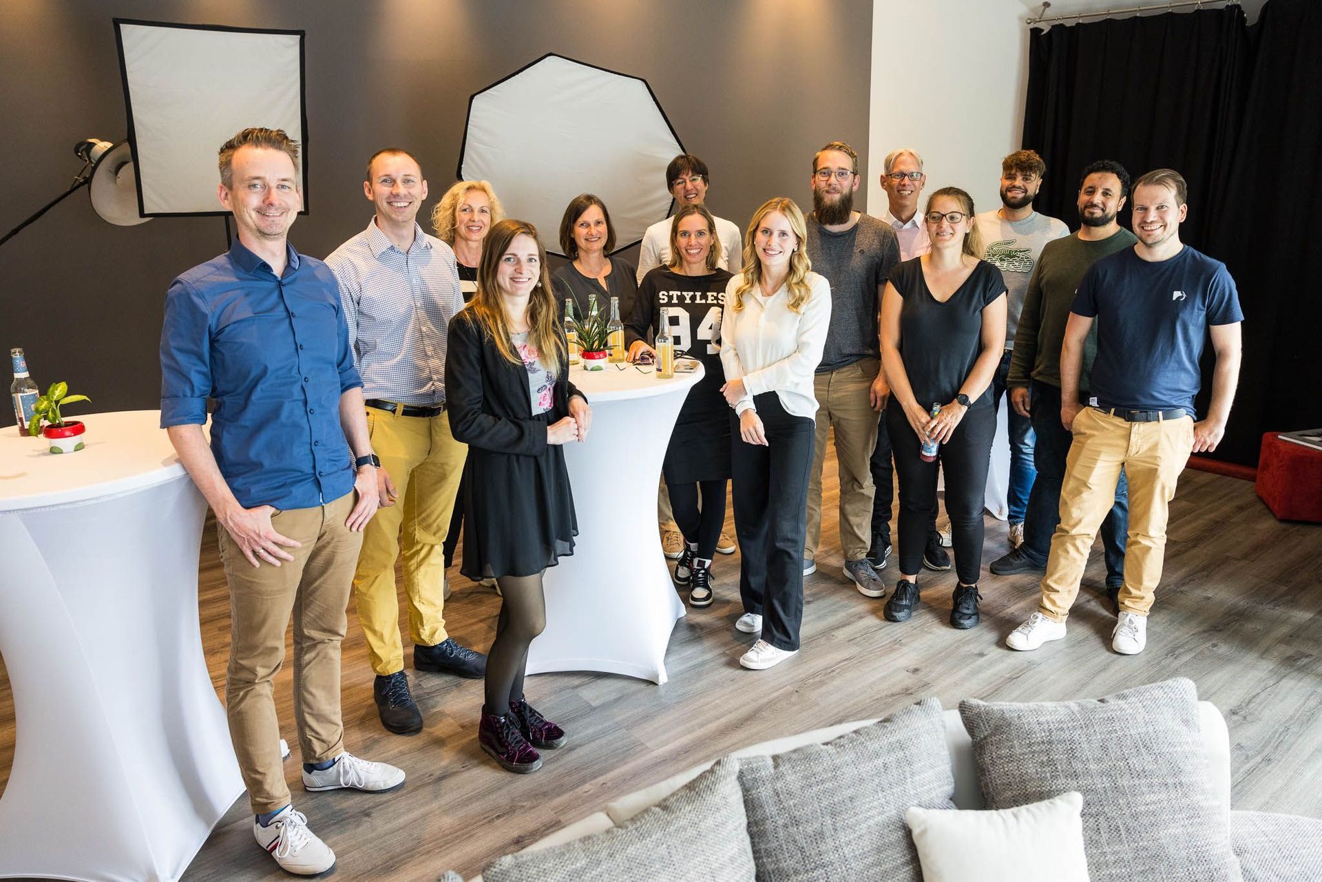 14 Personen verschiedener Geschlechter stehen lächelnd in einem Raum. Im Hintergrund steht Studiobeleuchtung.