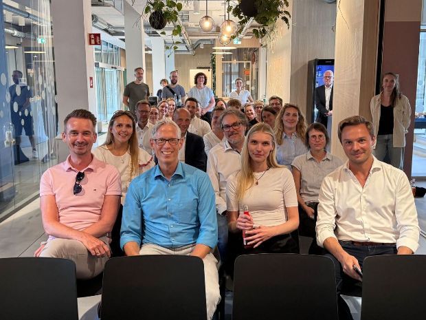 Eine Gruppe von Menschen sitzt in Reihen, der Kamera zugewandt, in einem hellen, modernen Raum.