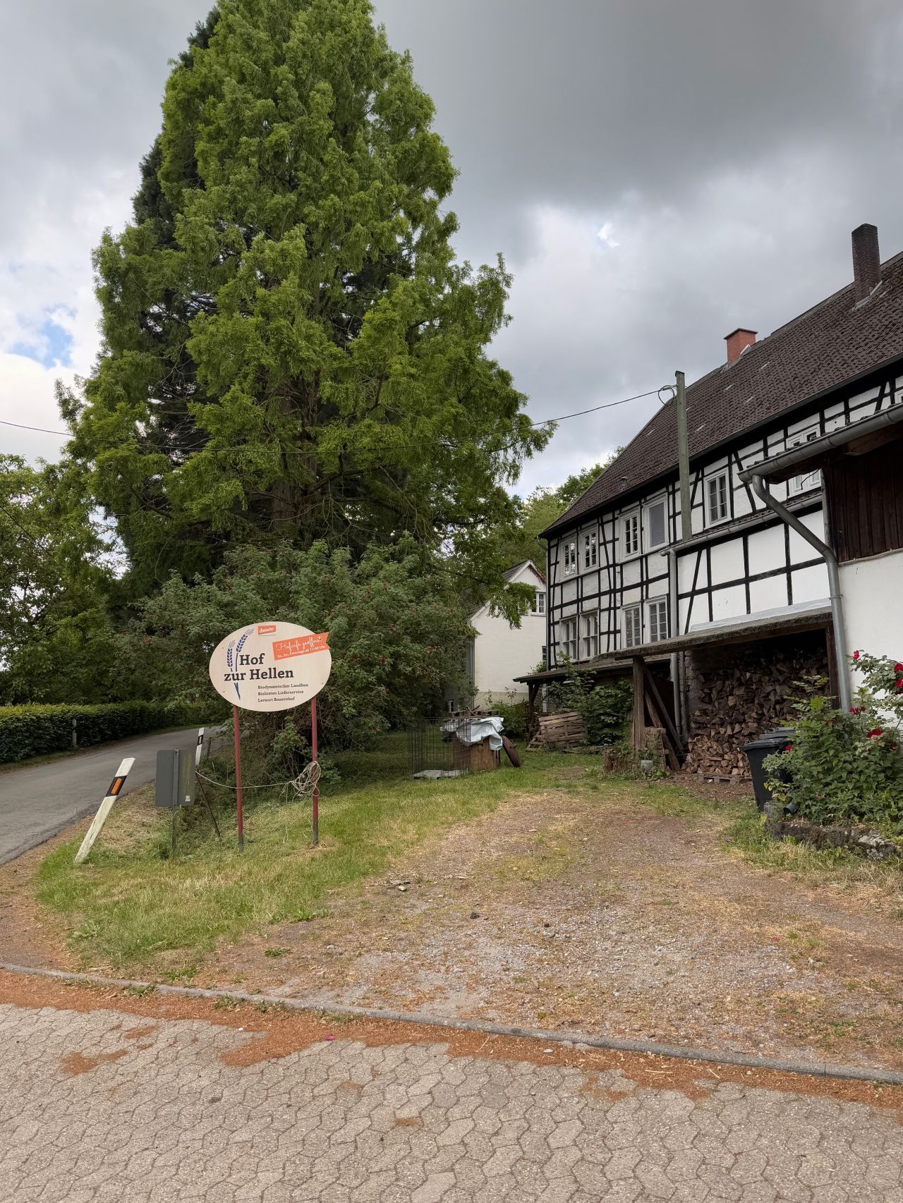 Einfahrt zum Biohof zur Hellen im Windrather Tal. Ein Schild und ein Fachwerkhaus sowie Bäume und ein bewölkter Himmel sind zu sehen.