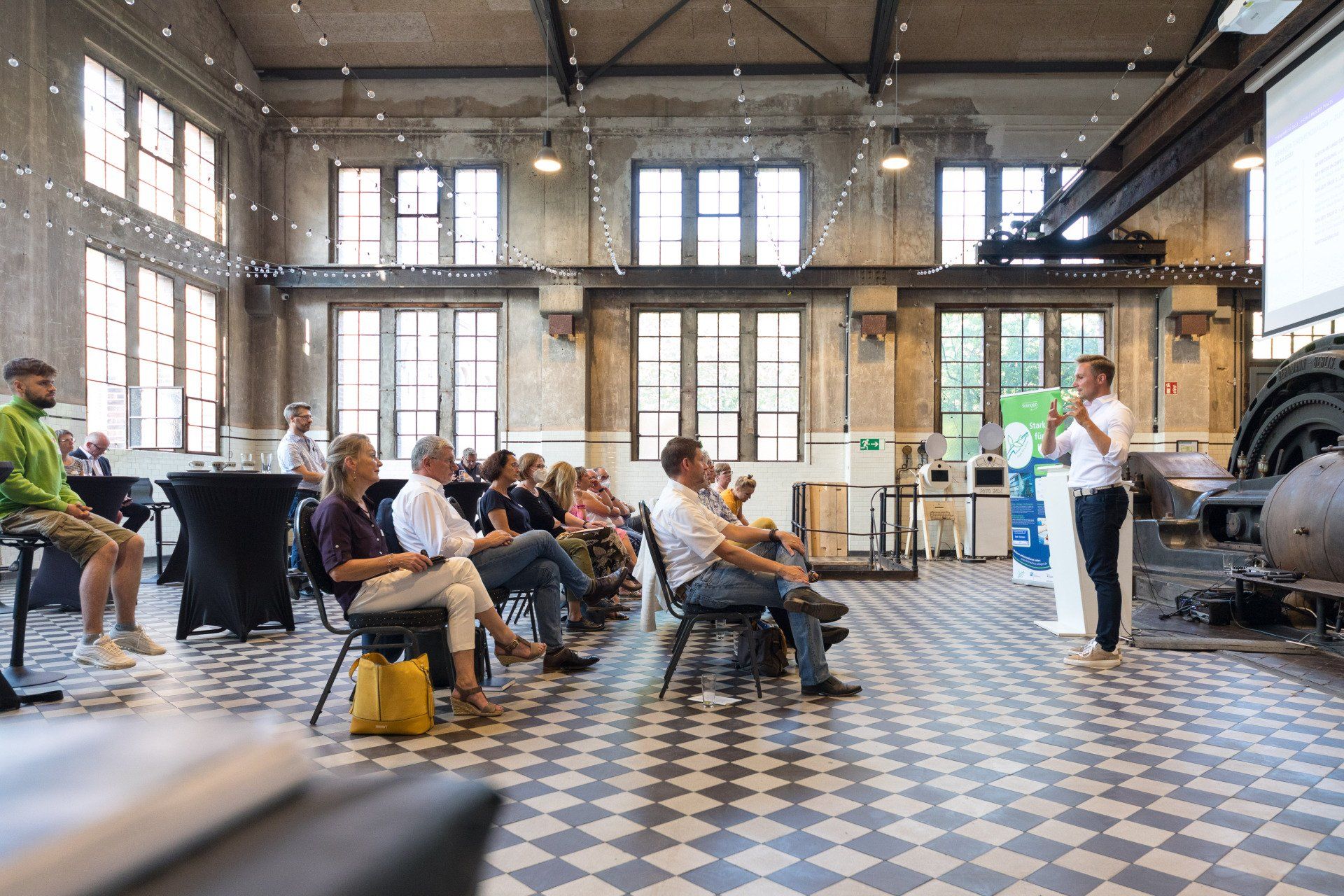 Ein Redner spricht zu einem Publikum verschiedener Geschlechter in einem industriellen Raum mit hohen Decken und Fenstern.