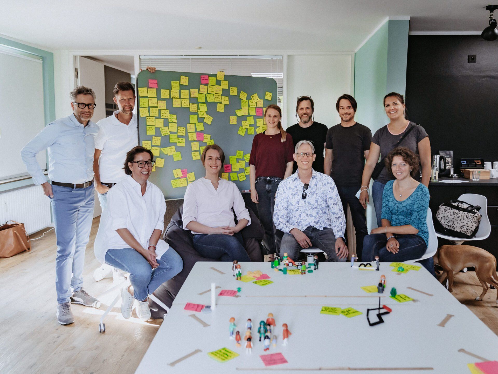 Eine Gruppe von zehn Personen sitzt bzw. steht lächelnd um einen Tisch mit kleinen Figuren und Post-it-Notizen herum.