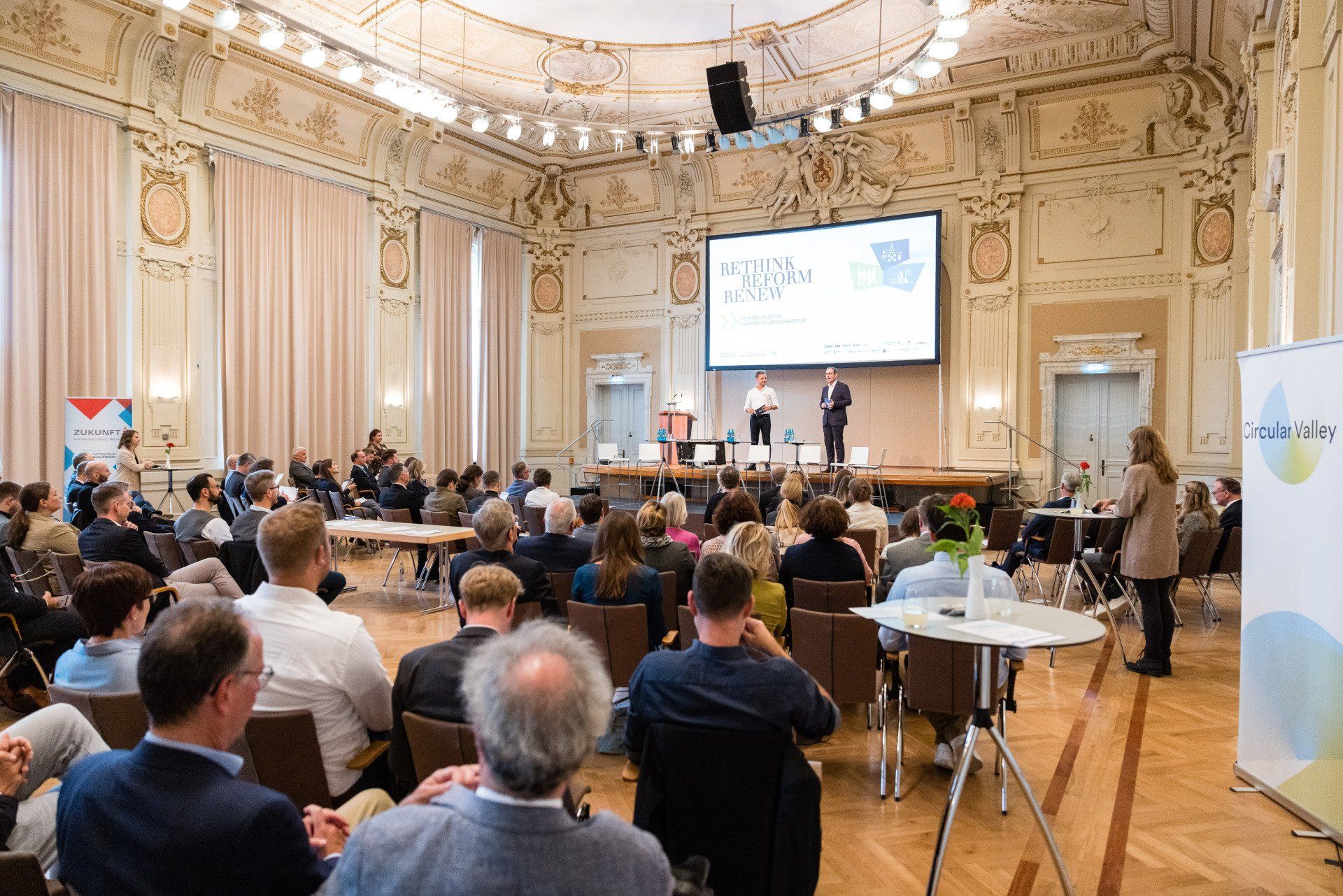 Eleganter Konferenzsaal mit hohen Decken und Ornamenten. Die Teilnehmer sitzen und beobachten zwei Redner auf der Bühne.
