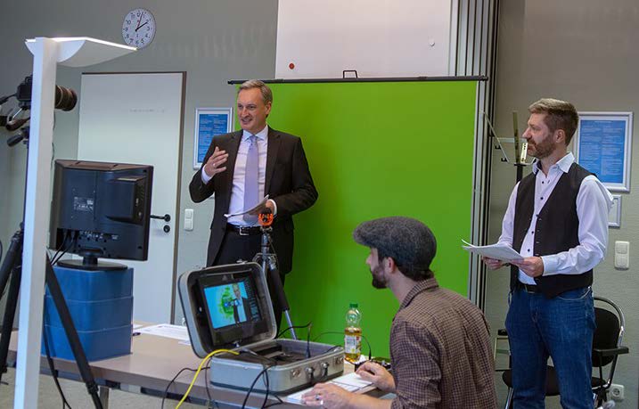 Ein Mann im Anzug steht vor einem Greenscreen und wird filmen. Zwei weitere Personen befinden sich neben dem Greenscreen.
