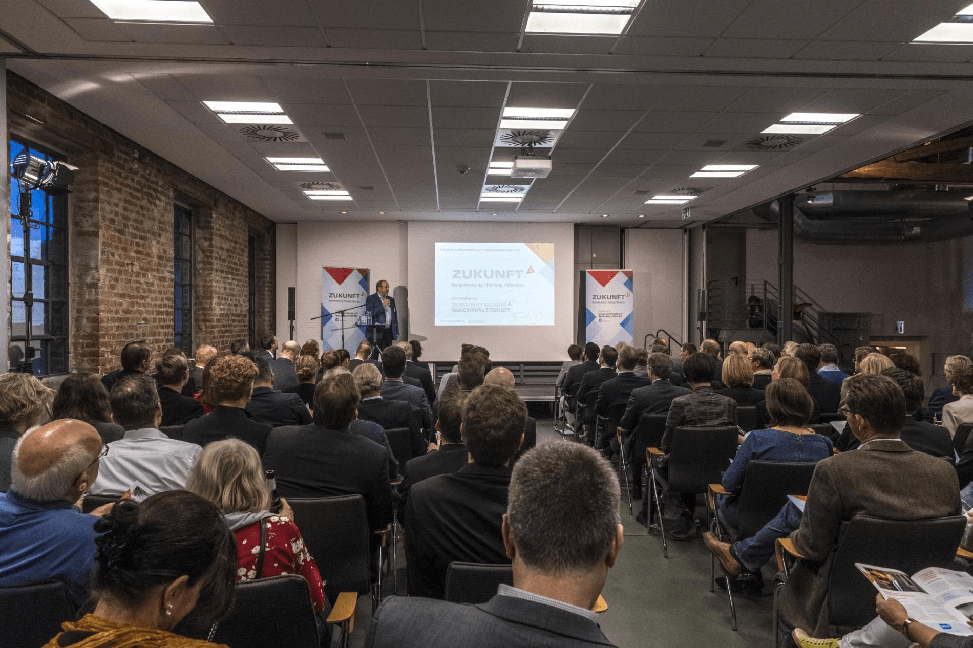 Personen in einem geräumigen Konferenzraum im Industriestil hören aufmerksam einem Redner auf einem Podium zu.