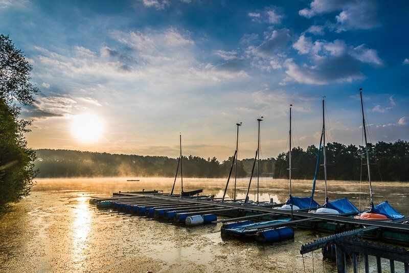 Die aufgehende Sonne beleuchtet einen See und einen Bootssteg mit kleinen Segelbooten.