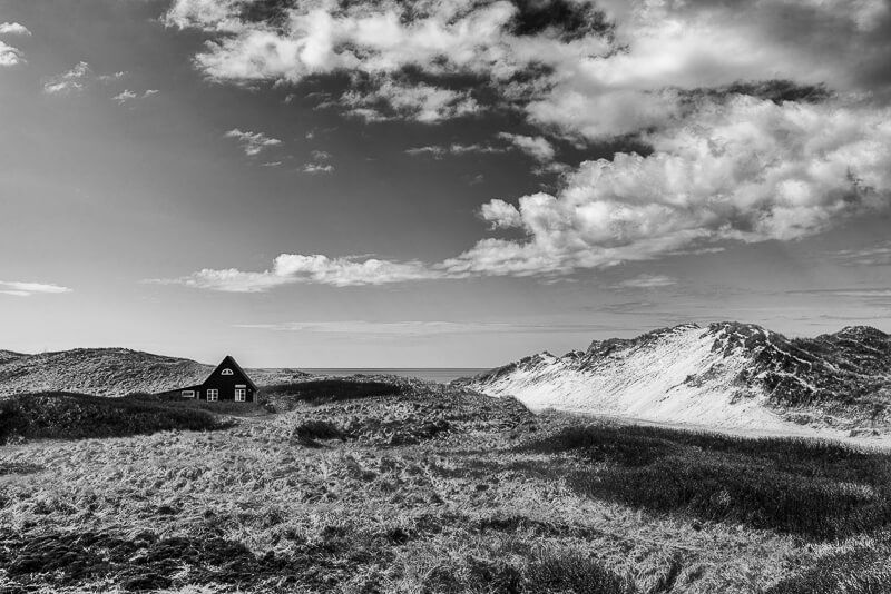 Eine schwarz-weiße Aufnahme mit Dünengras, links einem kleinen Haus und im Bildhintergrund ein Stückchen Ostsee.