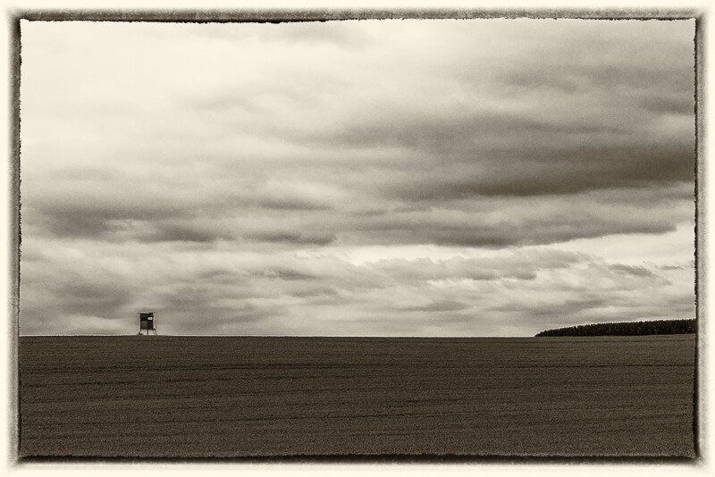Eine sepiagetönte Aufnahme von einem Feld mit bewölktem Himmel und in der linken Bildhälfte ein Hochsitz.