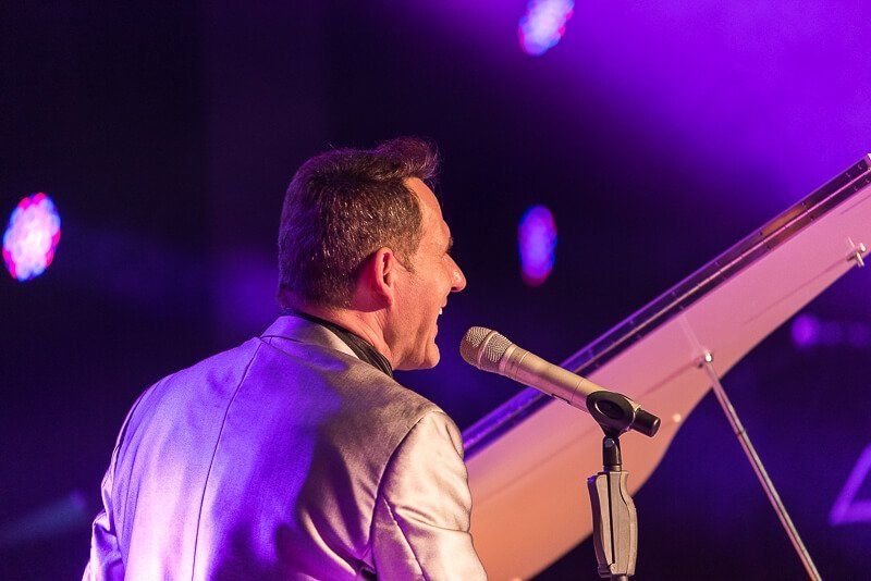 Ein singender Klavierspieler in einem silbernen Sakko seitlich von hinten fotografiert.