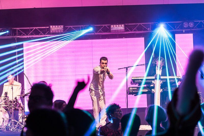 Ein Sänger auf einer Bühne zeigt ins Publikum und singt dabei. Links und rechts sind blaue Laserstrahlen zu sehen.