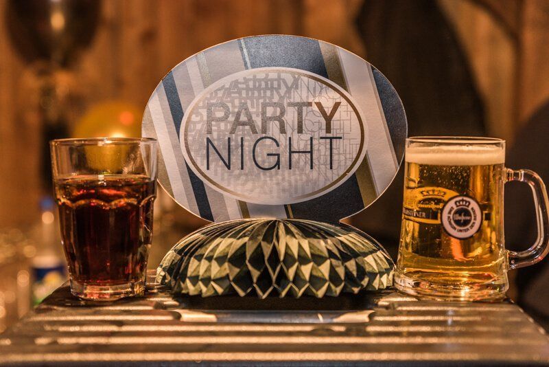 Ein Schild mit der Aufschrift Party Night. Rechts daneben ein volles Bierglas, links daneben ein volles Glas Cola.