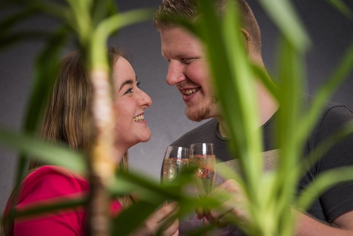 Portrait von einem jungen Paar durch eine Palme fotografiert. Sie stoßen mit einem Sektglas an und lächeln sich an.