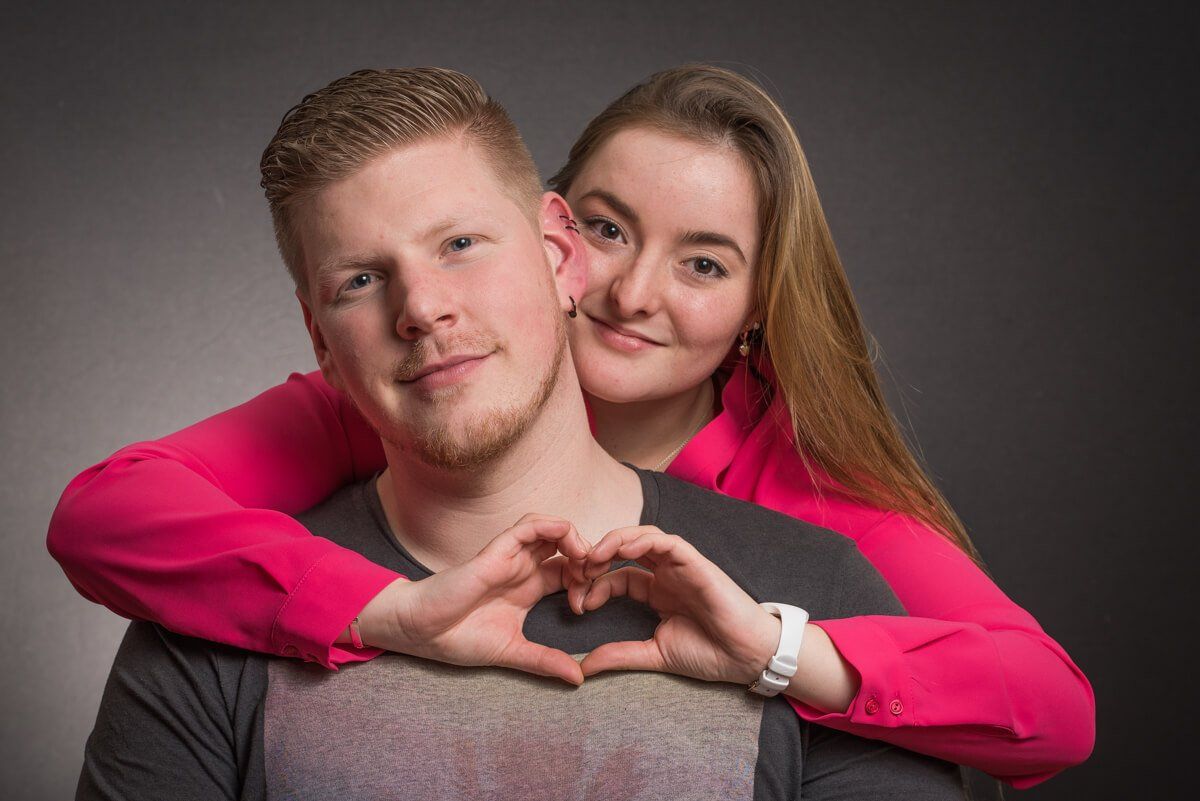 Portrait von einem jungen Paar. Sie steht hinter ihm und formt ihre Finger vor seiner Brust zu einem Herz.