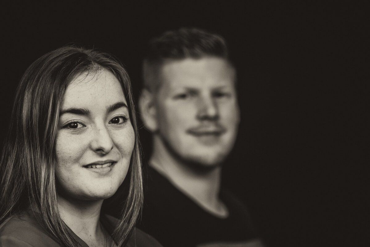 Portrait in schwarz-weiß von einem jungen Paar. Sie steht leicht lächeld im Vordergrund, er in leichter Unschärfe im Hintergrund.
