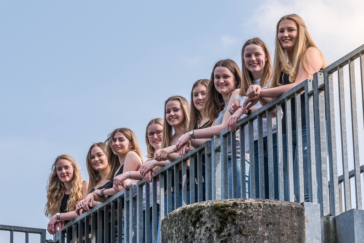 Neun junge Frauen, die nebeneinander an einem Geländer stehen und leicht von unten fotografiert wurden.