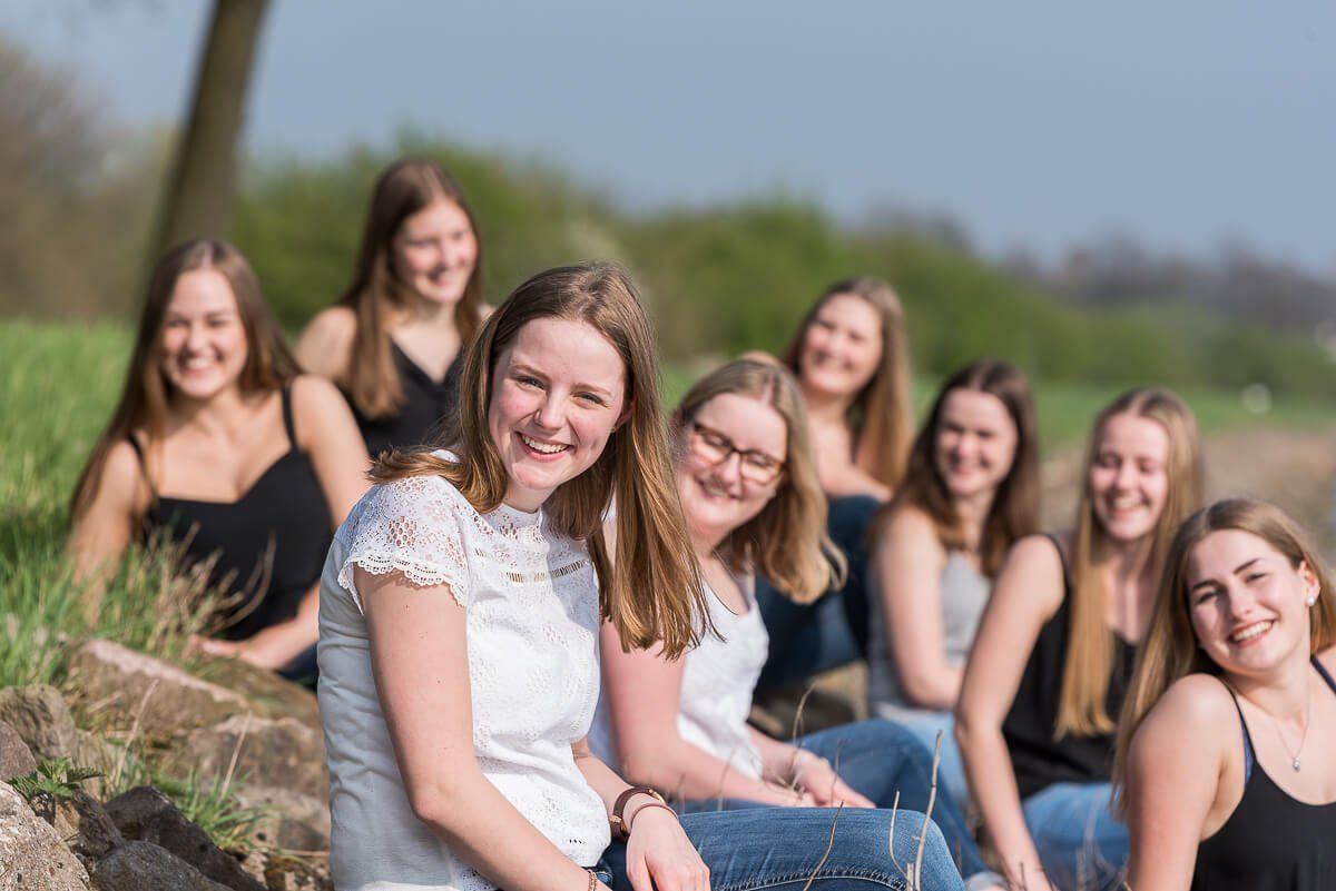 Neun junge Frauen, die in der Sonne sitzen. Eine davon ist in der Schärfe im Vordergrund, die anderen sind in der Unschärfe im Hintergrund.
