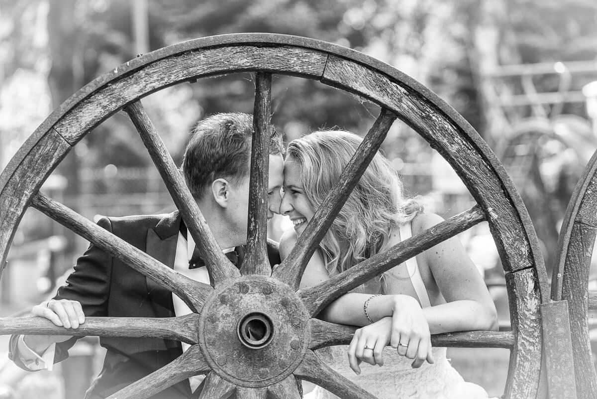 Ein Hochzeitspaar in weißem Kleid und schwarzem Smoking hockt hinter einem alten Wagenrad aus Holz. Sie lehnen Stirn an Stirn und lächeln sich an.